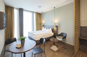 Una habitación de hotel con una cama, dos sillas, una mesita, un televisor y una planta.