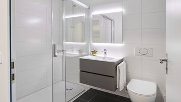 Un baño moderno con cabina de ducha de vidrio, un lavamanos blanco con detalle dorado y un inodoro blanco.