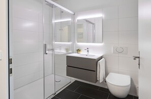 Un baño moderno con cabina de ducha de vidrio, un lavamanos blanco con detalle dorado y un inodoro blanco.