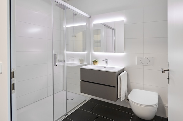 A modern bathroom with a glass shower enclosure, a white sink with a gold accent, and a white toilet. - Lumen Apartments (Schenkon)