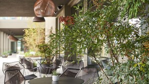 An outdoor seating area with round tables and black wicker chairs, surrounded by greenery and hanging pendant lights. - Lumen Apartments (Schenkon)