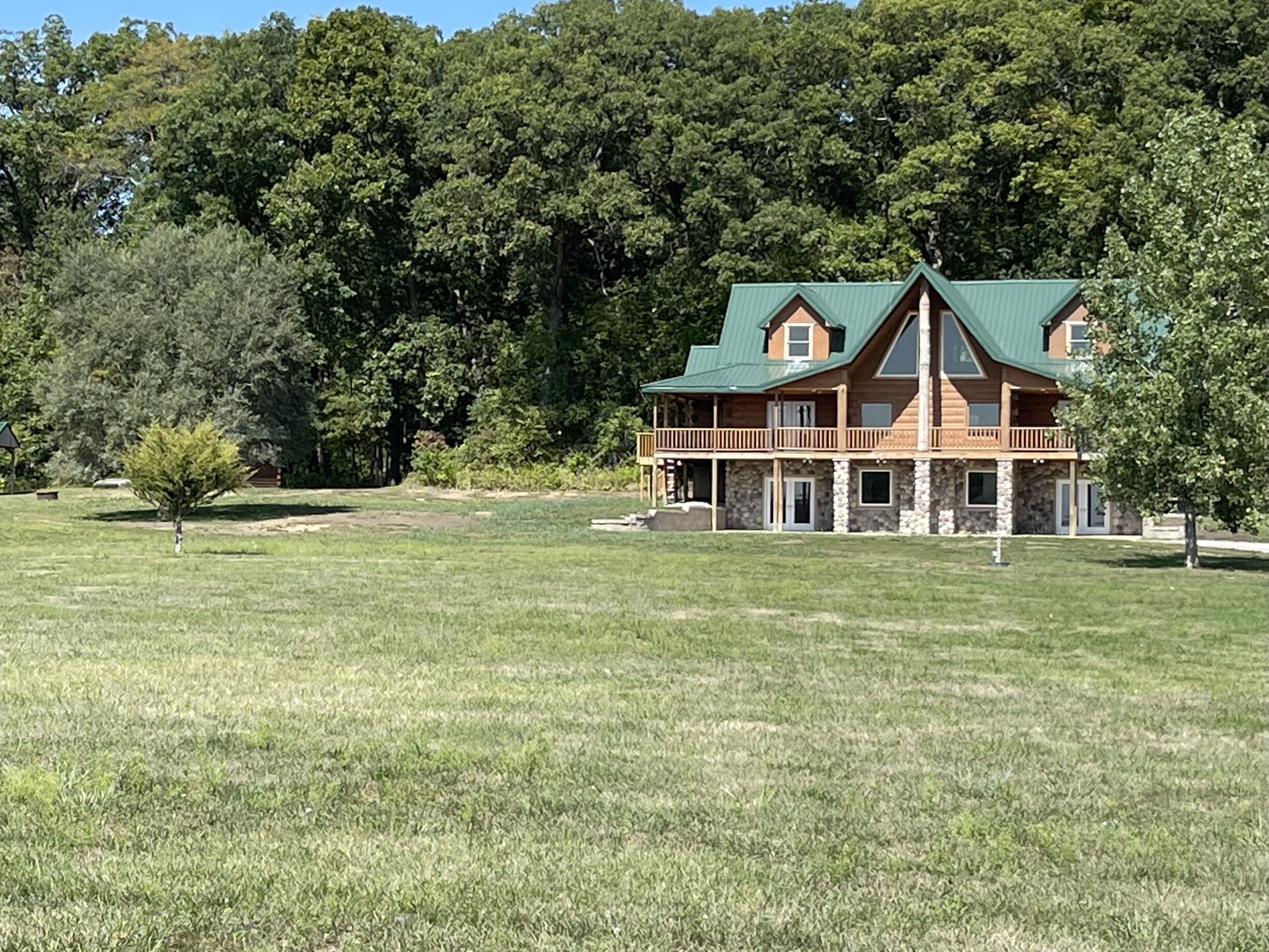 A large, two-story log cabin with a green roof, surrounded by a grassy field and dense forest.