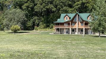 A large, two-story log cabin with a green roof, surrounded by a grassy field and dense forest.
