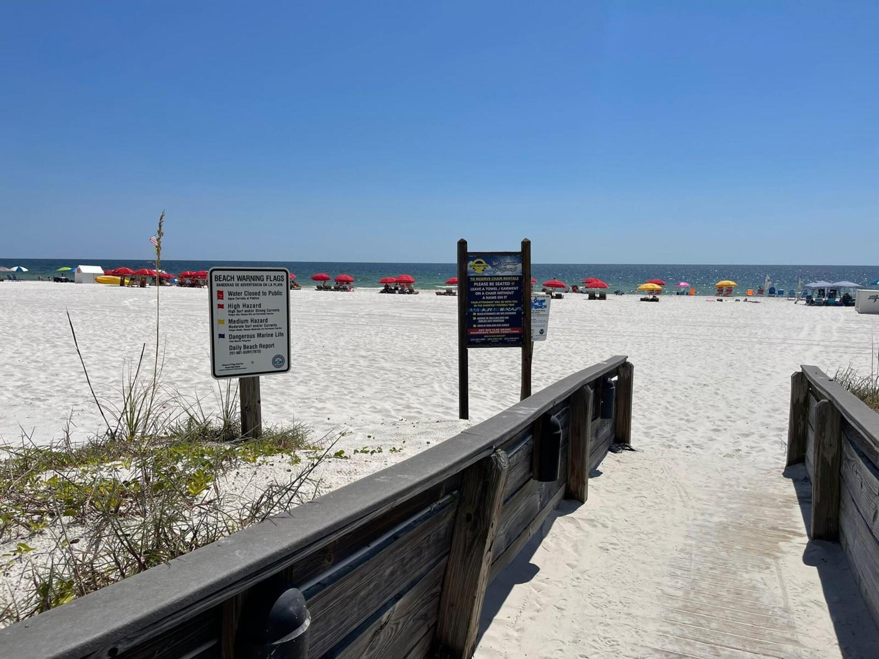 A beach with a wooden boardwalk leading to the water, beach warning flags, and beach chairs under umbrellas.