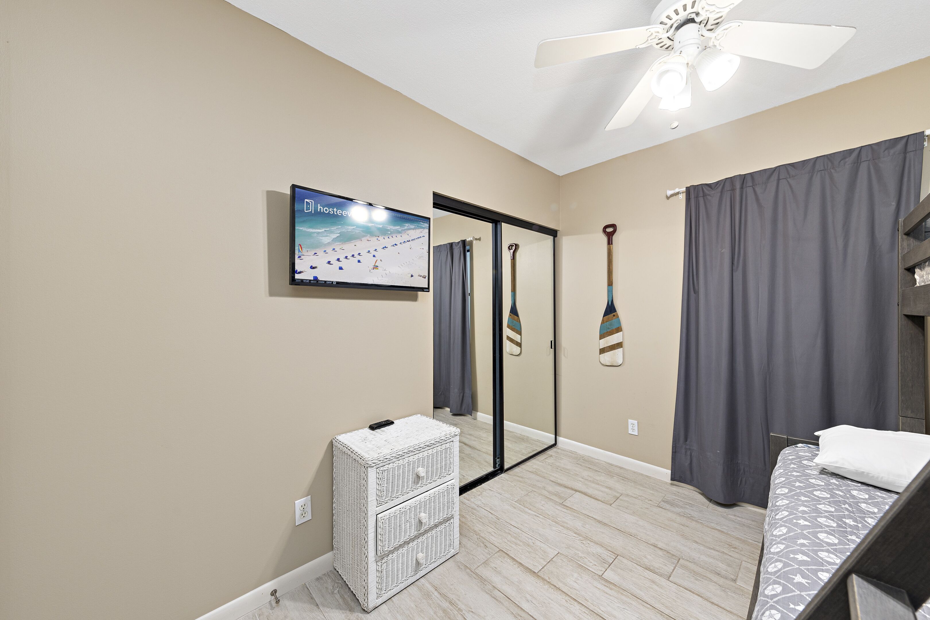 A bedroom with a bed, a television, a mirror, a ceiling fan, and a small white chest of drawers.