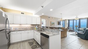 A modern kitchen with white cabinets, a granite countertop, and a dishwasher. There is a dining area with a large window overlooking the ocean. - Shoalwater 804 (Orange Beach)