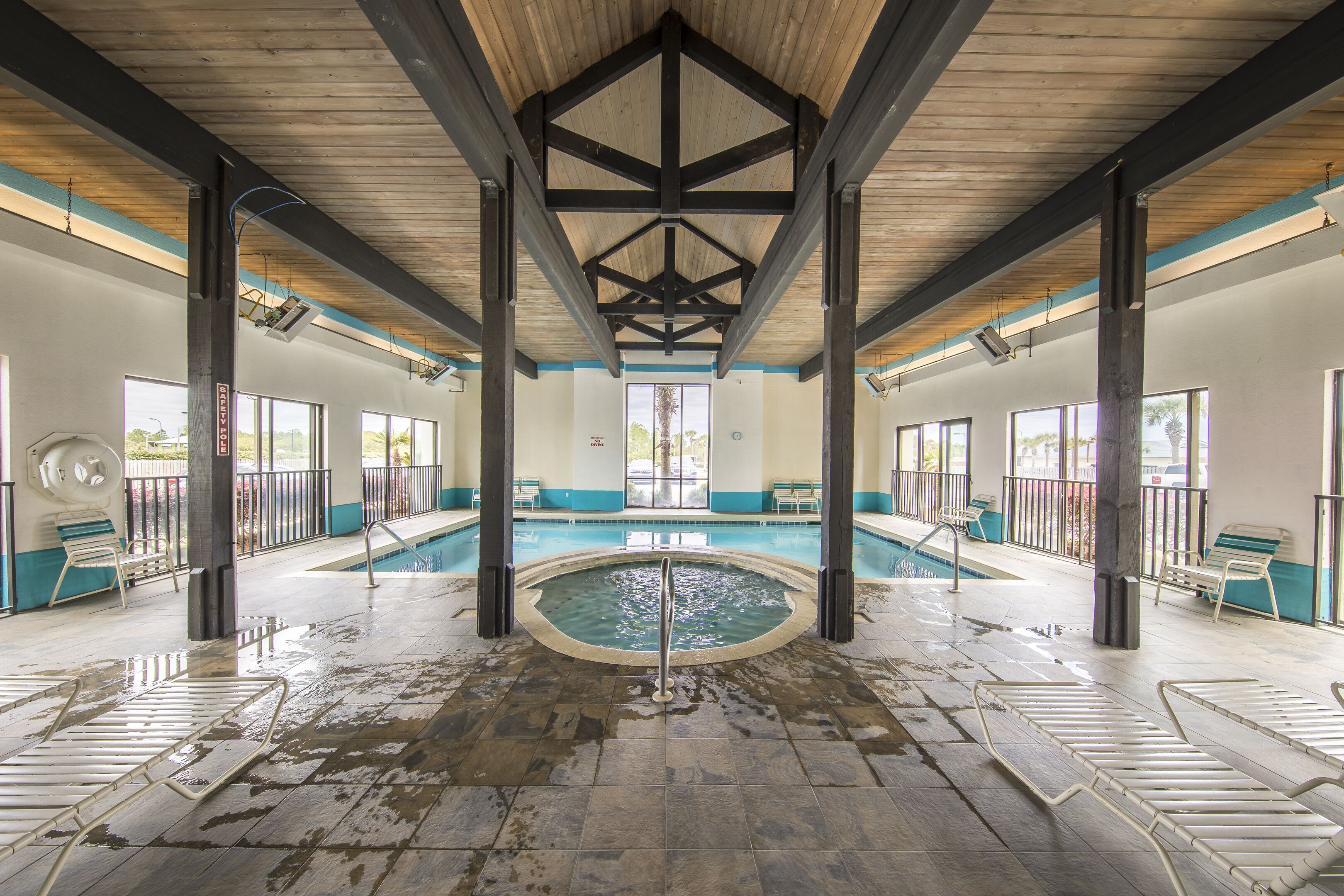 An indoor swimming pool with a wooden ceiling and tiled floor, surrounded by lounge chairs and a view of the outdoors.