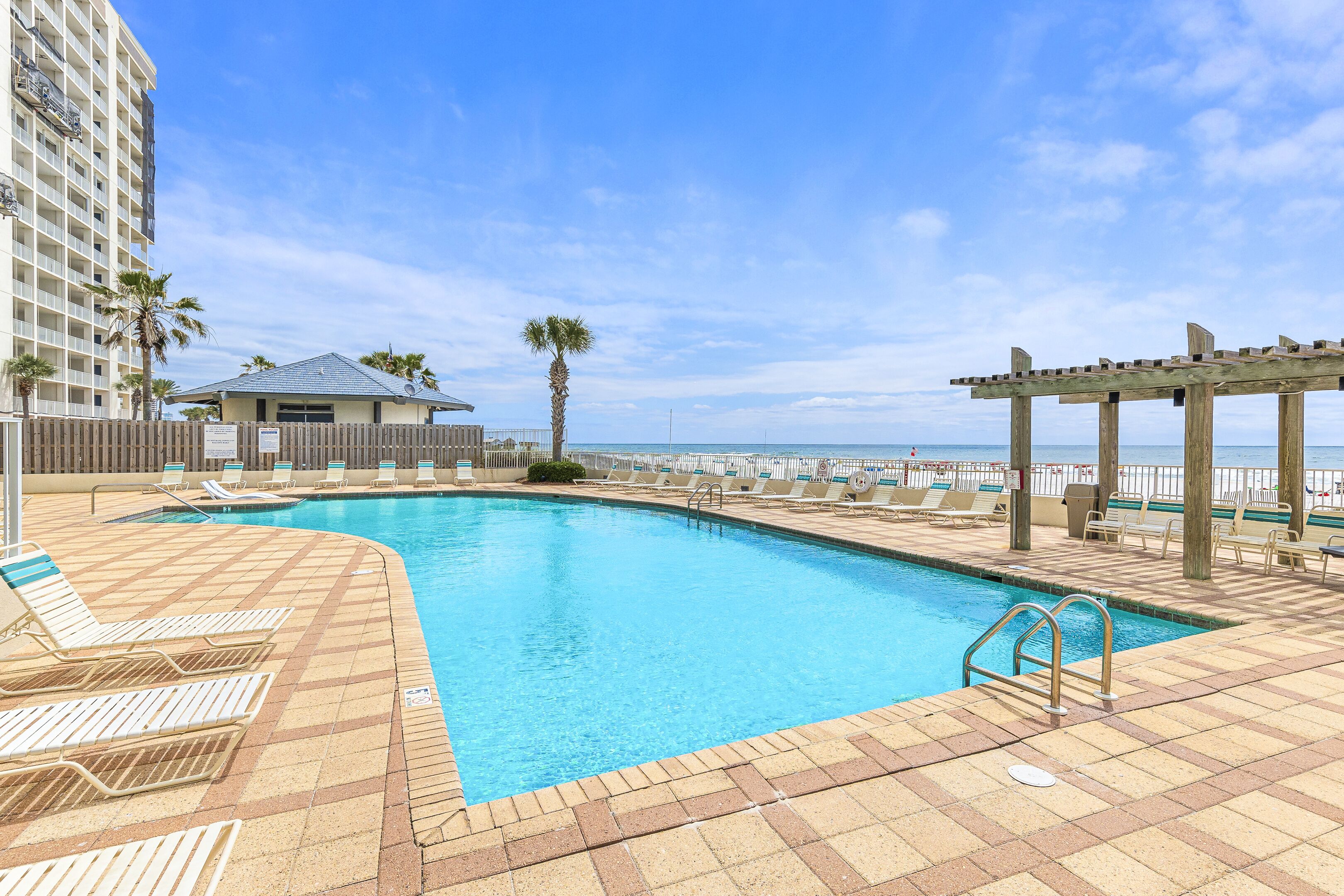 A swimming pool with lounge chairs and a pergola, surrounded by a high-rise building and a beach view.
