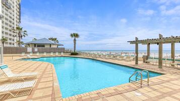 A swimming pool with lounge chairs and a pergola, surrounded by a high-rise building and a beach view.