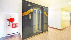A hallway with a fire extinguisher, a fire alarm, and a glass elevator door with a yellow accent. - PPH Living Tricolours (Coimbatore)