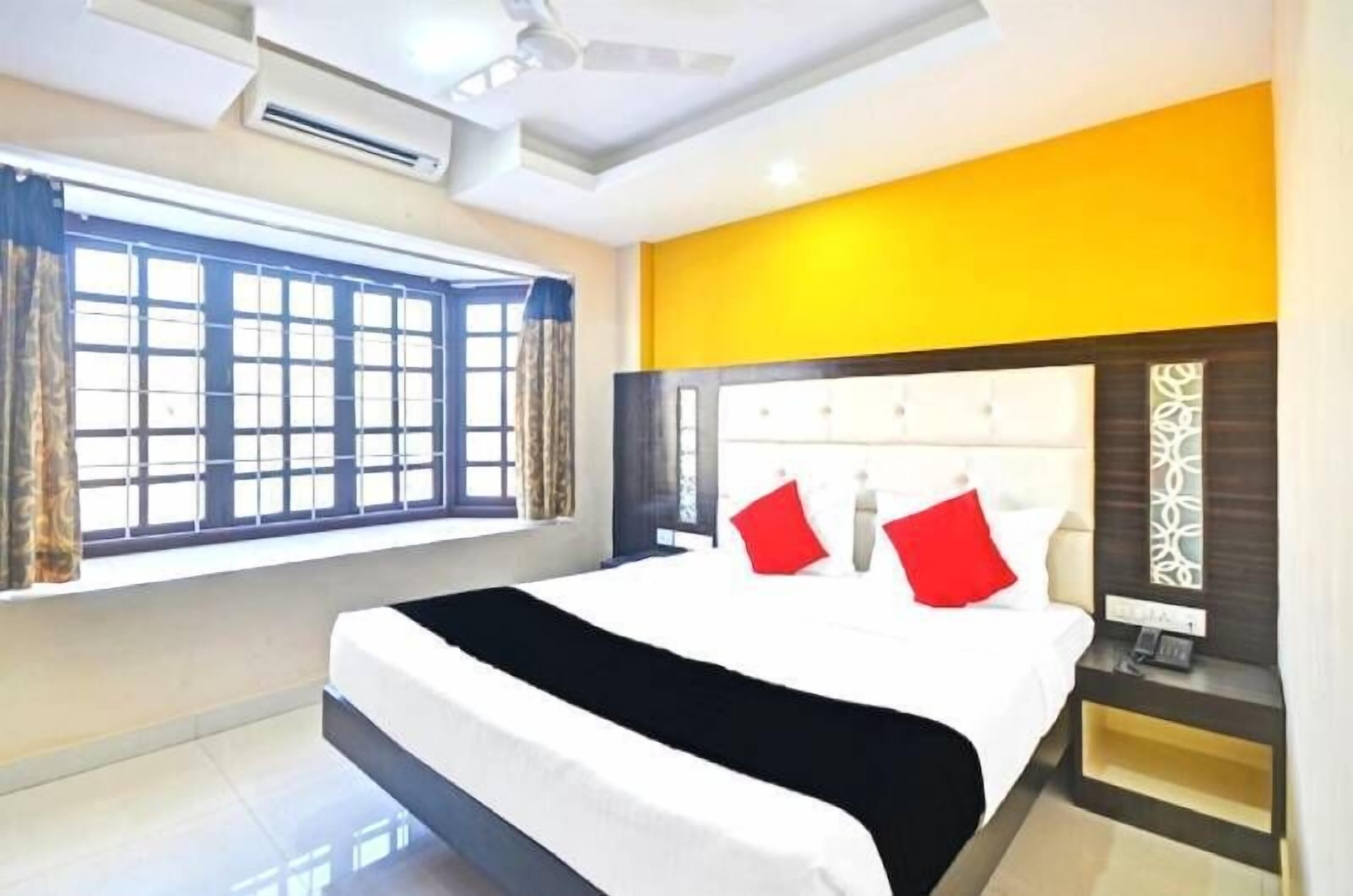 A hotel room with a large bed, a bedside table with a lamp, a window with curtains, and an air conditioning unit.