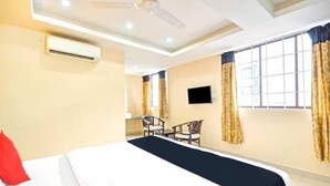 A hotel room with a bed, a desk, two chairs, a TV, and large windows with curtains. - PPH Living Tricolours (Coimbatore)