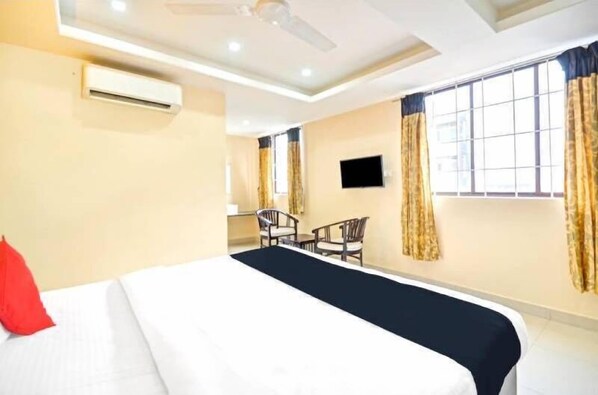 Quarto de hotel com cama, escrivaninha, duas cadeiras, televisão e janelas amplas com cortinas.