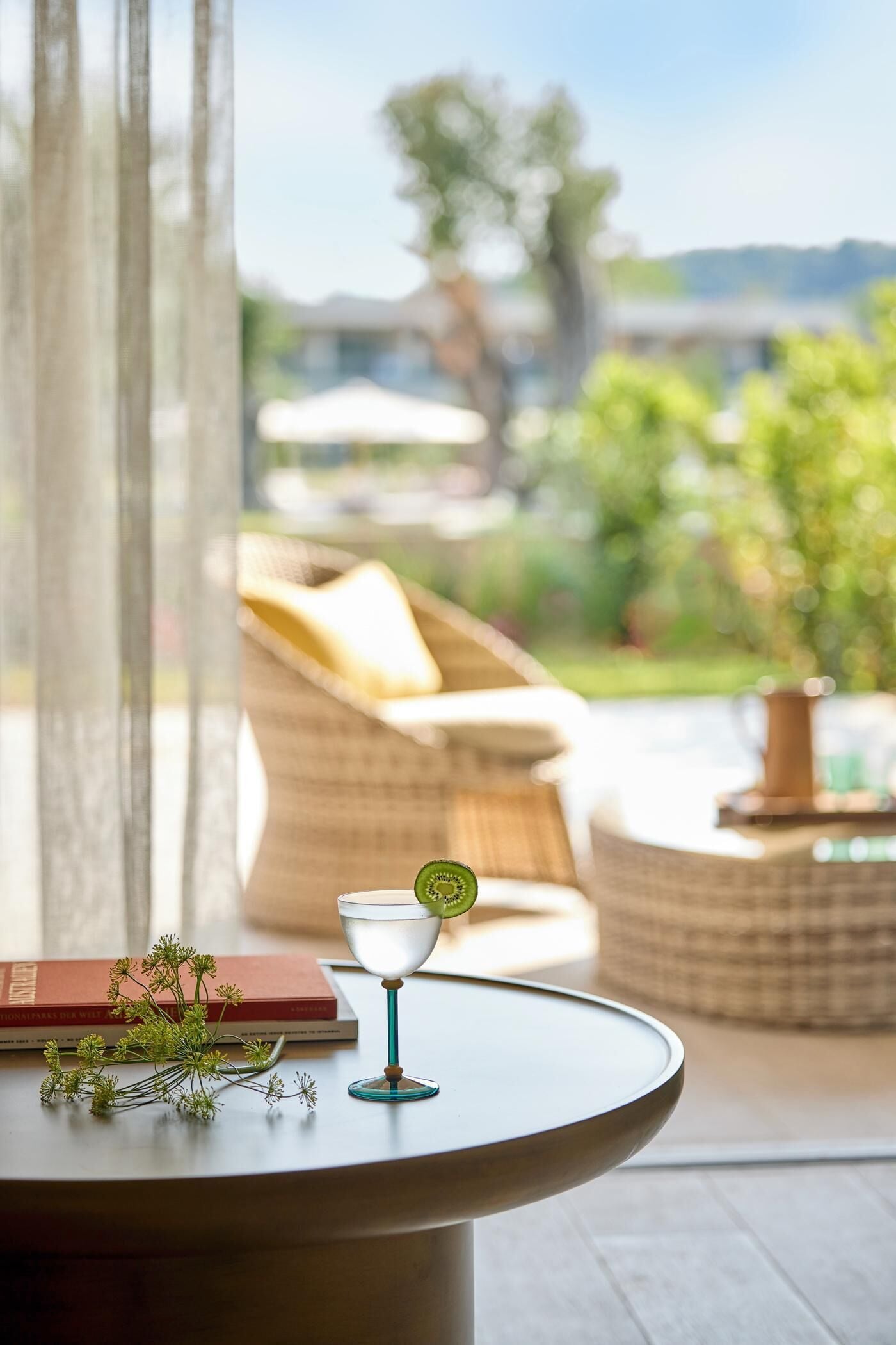 Una mesa redonda con un libro y un vaso, una silla de mimbre y vistas a un jardín y edificios.