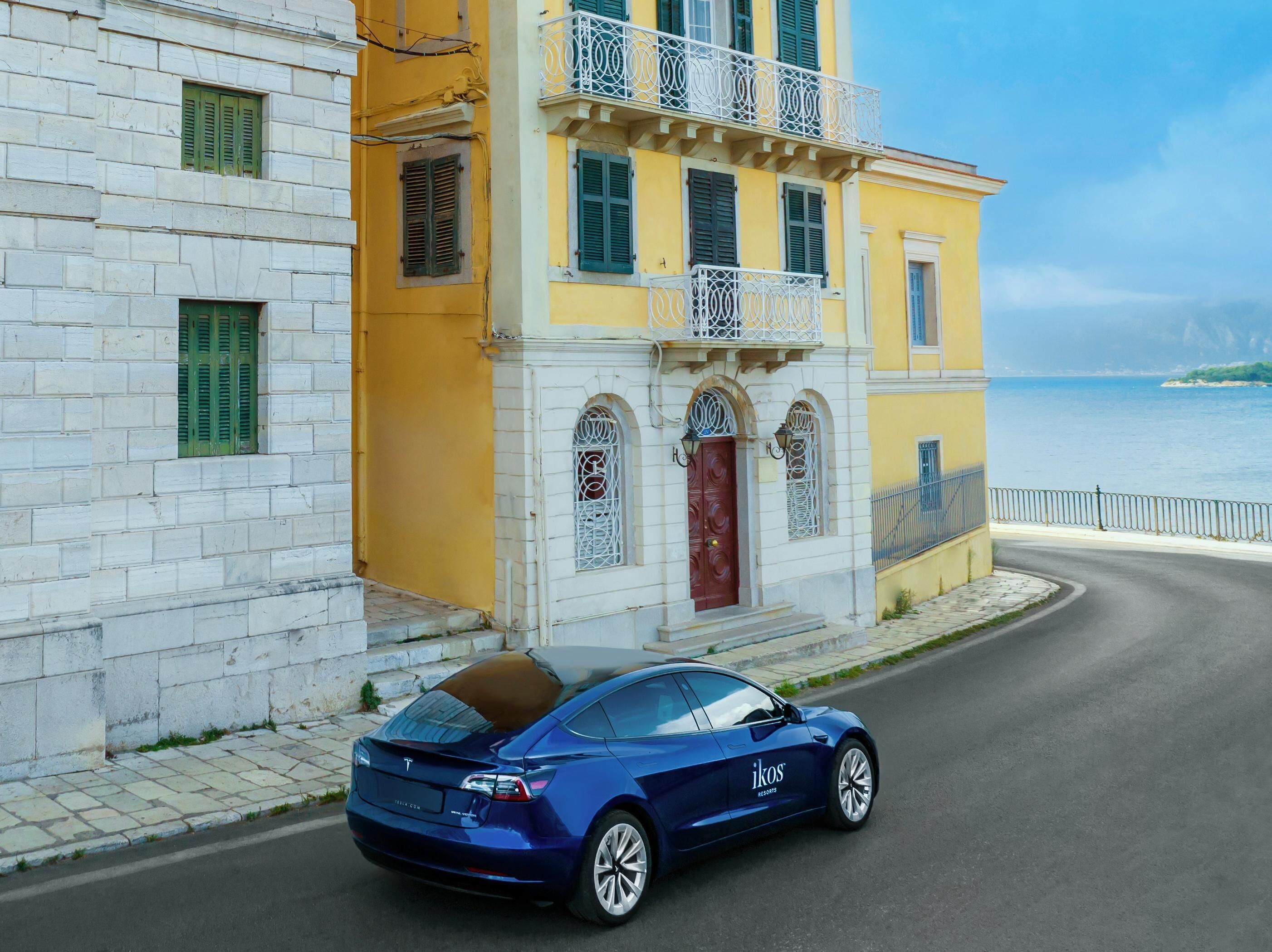 A blue Tesla parked in front of a yellow building with a white facade and green shutters.