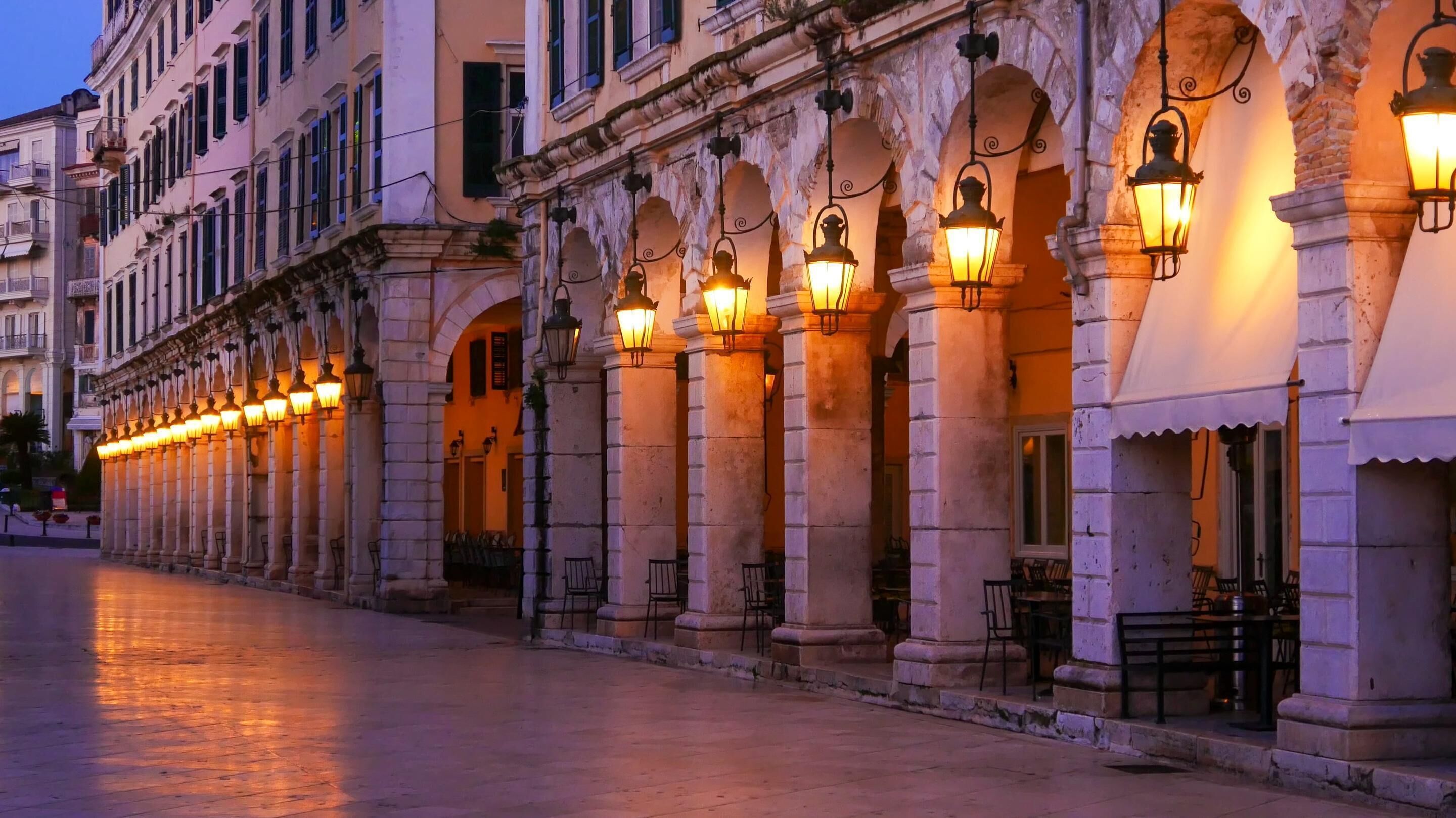 Un edificio histórico con una fila de arcos y columnas, asientos al aire libre e iluminación tradicional.