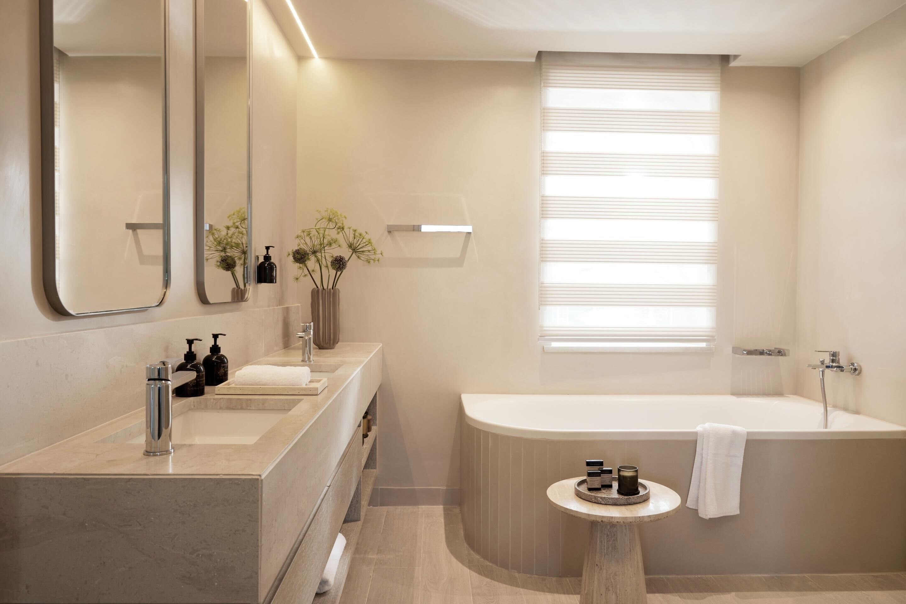 A modern bathroom with a large mirror, two sinks, a bathtub, and a small round table with a towel.