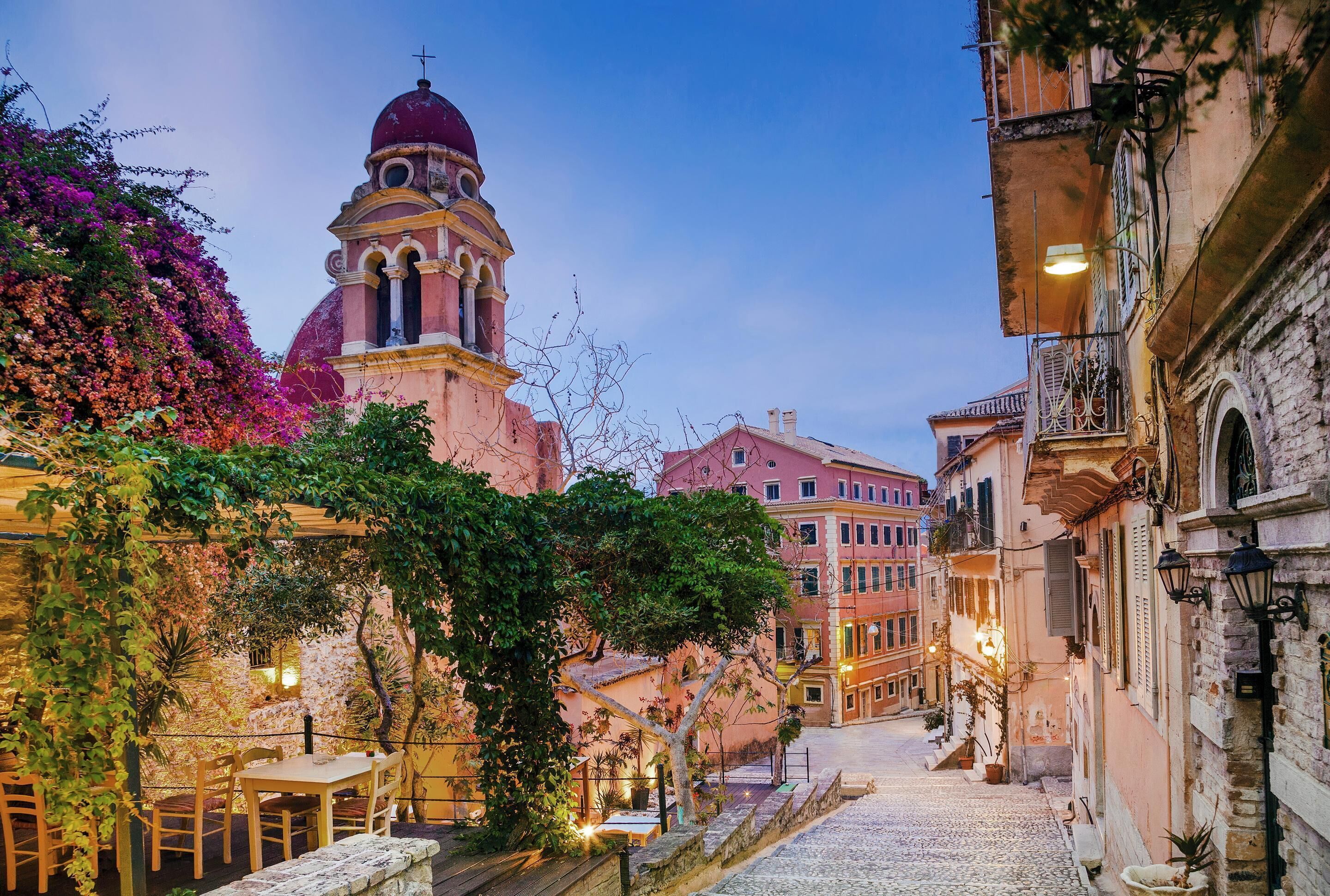 Una strada acciottolata fiancheggiata da edifici, con un campanile e tavolini e sedie all'aperto.
