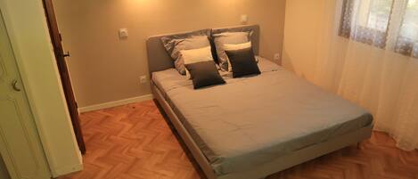 A bedroom with a bed, wooden flooring, and a window with curtains.