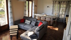 A living room with a grey sectional sofa, a small wooden table, and a dining area with a white table and chairs. - Gîte en Pleine Nature (Valaurie)