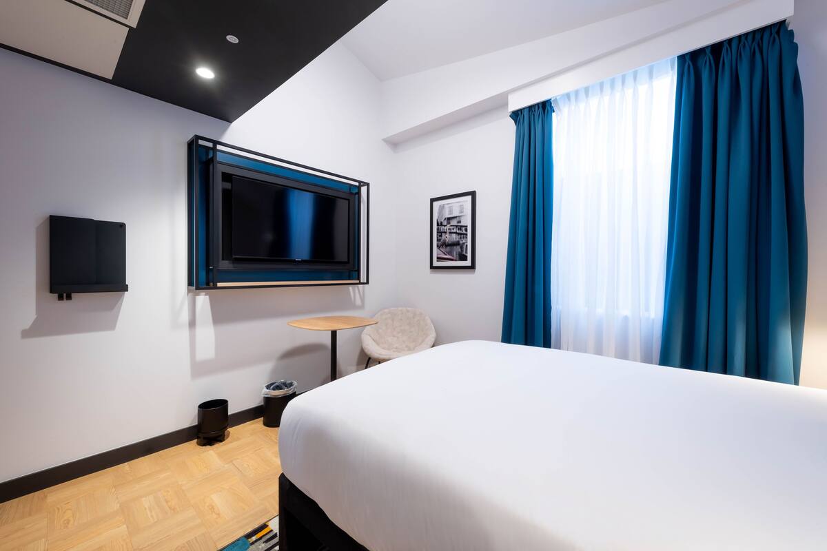 A modern hotel room with a large bed, a flat-screen TV mounted on the wall, a small round table, and a chair.
