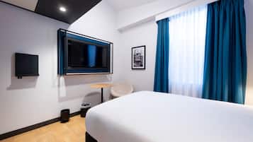 Habitación de hotel con cama, televisor montado en la pared, una mesita redonda, una silla y una ventana con cortinas azules.