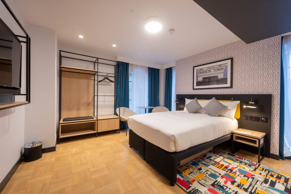 A modern hotel room with a bed, a desk, a chair, a wardrobe, and a patterned rug.