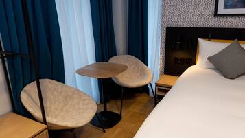 Habitación de hotel con cama, dos sillas, una mesita, una mesita de noche y una lámpara de pared.