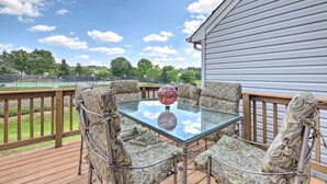 A deck with a glass table and patterned chairs, overlooking a tennis court and greenery. - Pond-View Deck + Pool Table: Woodbridge Haven! (Woodbridge)