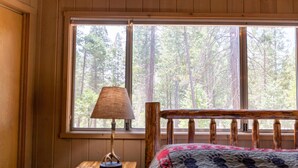Una cama de madera con cabecera, una mesita de noche con lámpara, y una ventana con vista al bosque.