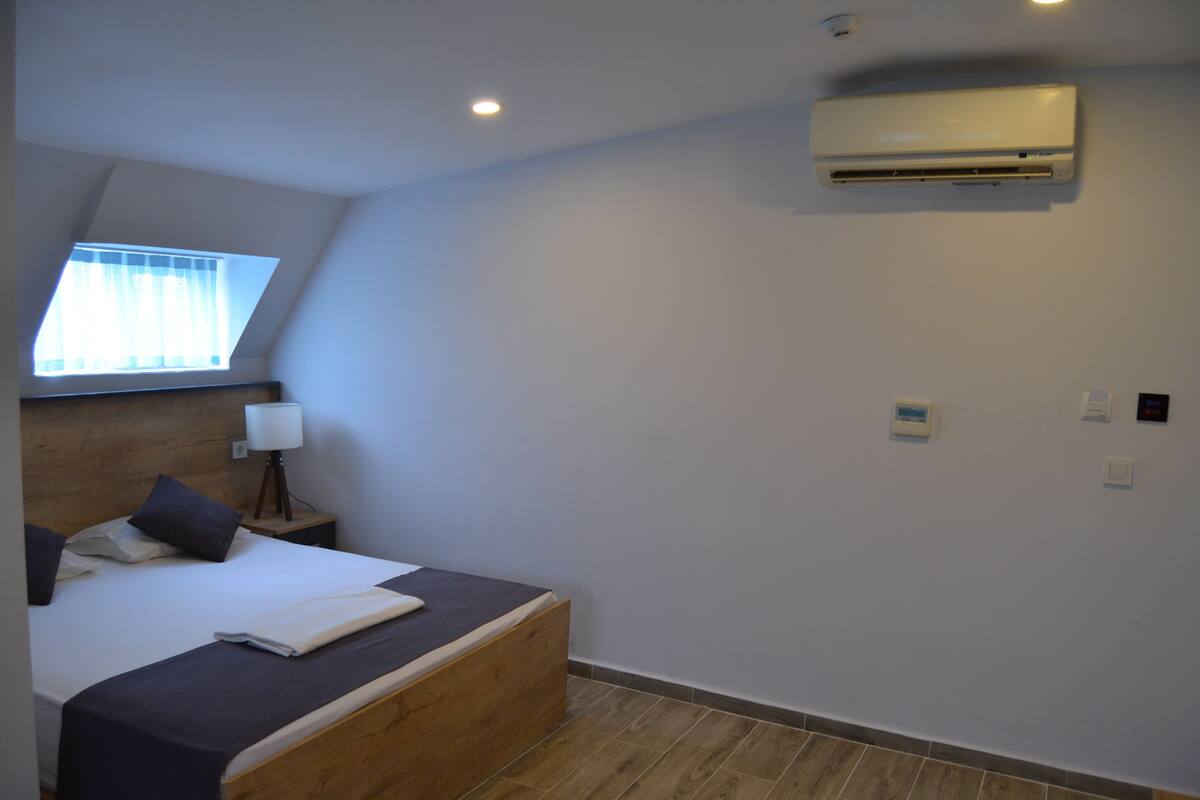 A bedroom with a bed, a nightstand, a lamp, a window with curtains, an air conditioner, and a wall-mounted control panel.