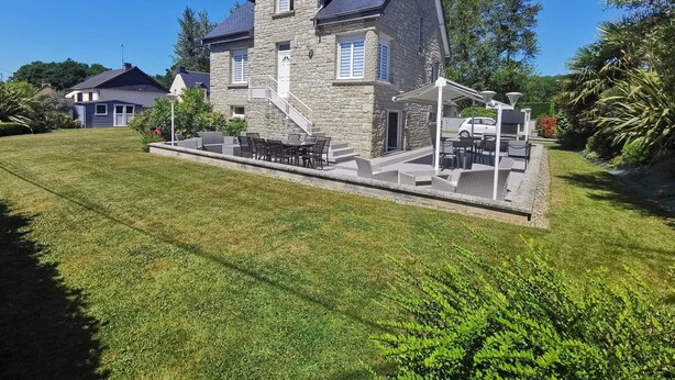 A stone house with a sloped roof, a covered patio with outdoor furniture, and a well-maintained lawn. - Loft Proche Plage (Pleslin-Trigavou)