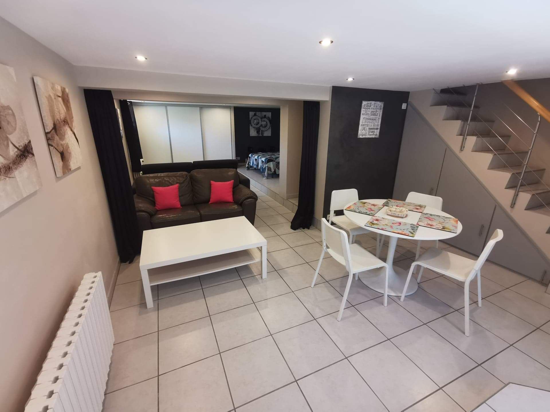 A modern living room with a sofa, coffee table, and dining table. There is a staircase to the right and a view of the outside.
