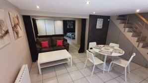 A modern living room with a sofa, coffee table, and dining table. There is a staircase to the right and a view of the outside. - Loft Proche Plage (Pleslin-Trigavou)