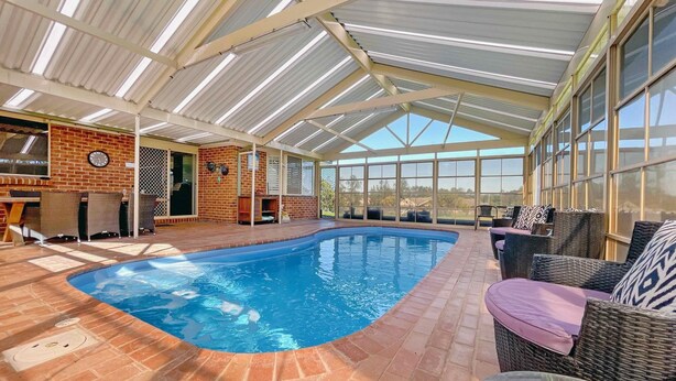 Una piscina interior con suelo de ladrillo, rodeada de mobiliario de mimbre y un techo de cristal.