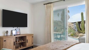 A bedroom with a bed, a TV mounted on the wall, a wooden cabinet with small plants, and a sliding glass door leading to an outdoor area with cacti.