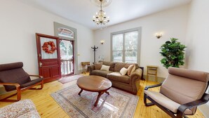 A living room with a wooden floor, a brown sofa set, a round coffee table, a brown armchair, a red door with a wreath, and a large green plant. - Retha Victorian - Gunnison (Gunnison)