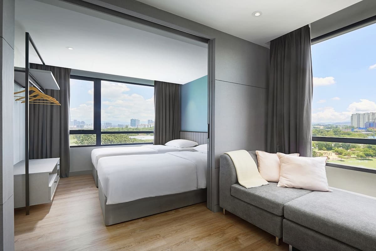 A modern hotel room with two beds, a sofa, a desk, and large windows offering a city view.