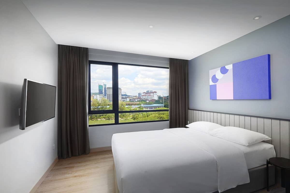 A modern hotel room with a large bed, a flat-screen TV, a city view through the window, and a contemporary wall art piece.