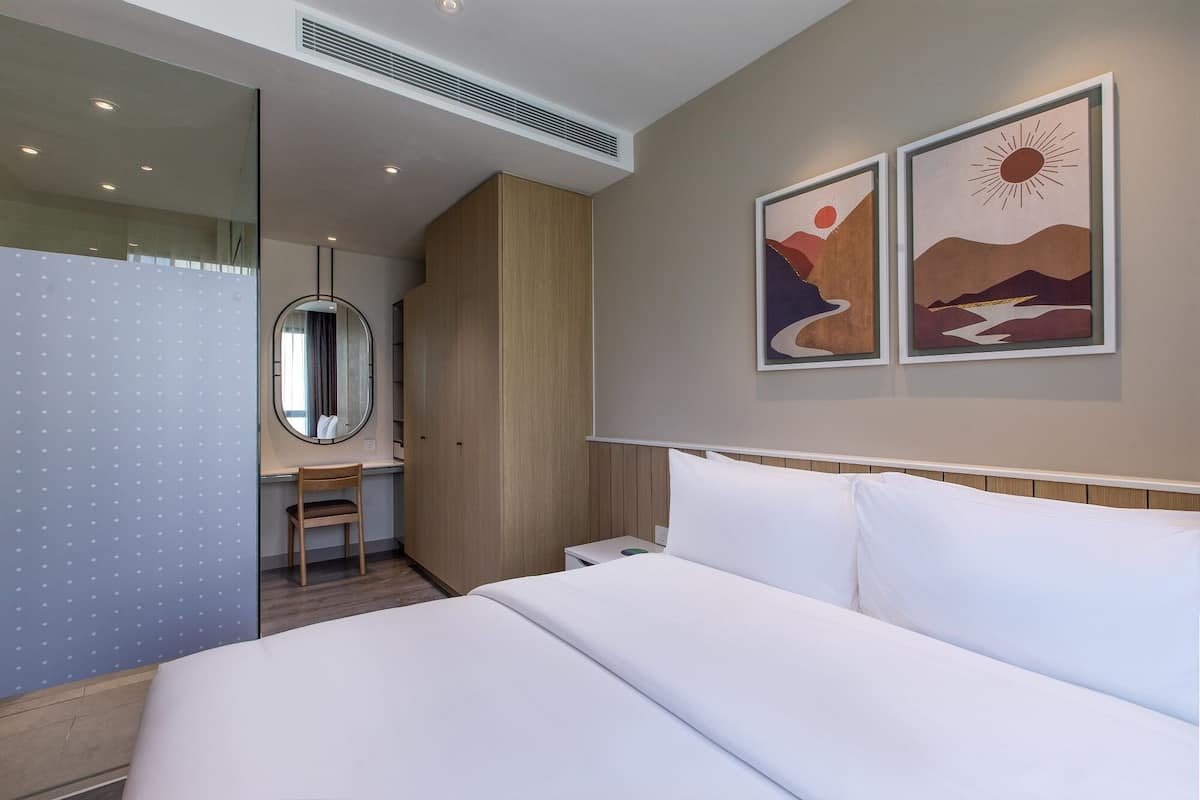 A hotel room with a bed, a desk, a chair, a wardrobe, a mirror, and two wall-mounted paintings.
