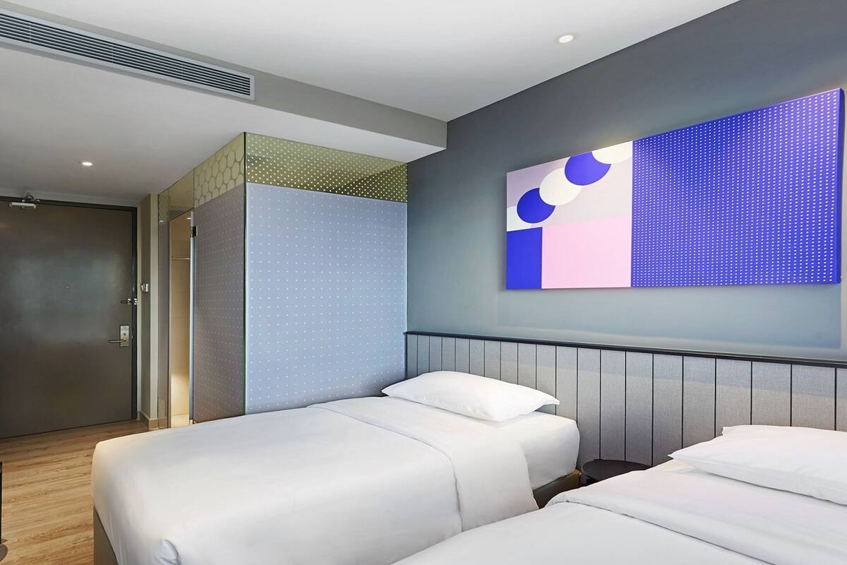 A hotel room with two beds, a desk with a chair, a wardrobe, and a large abstract painting on the wall.