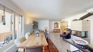A spacious living area with a long wooden dining table, comfortable seating, a fireplace, and a large window providing natural light. - 3½ room apartment, on the second floor, with lift. (Samedan)