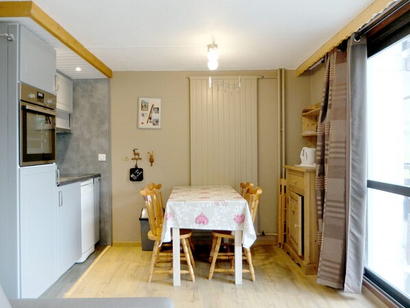 A compact kitchen with a dining area, wooden chairs, a table with a floral tablecloth, and a window with curtains.