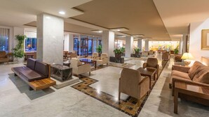 A spacious lobby with a modern design, featuring a central seating area with sofas and armchairs, a marble floor, and large windows providing natural light.
