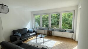 Ein modernes Wohnzimmer mit einem schwarzen Ledersofa, einem Couchtisch und großen Fenstern, die einen Blick auf Grünfläche bieten.