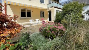 Ein zweistöckiges Haus mit einem Balkon, einer kleinen Terrasse mit Stühlen und einem Garten mit verschiedenen Pflanzen und Blumen.