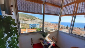 A balcony with a table and chairs, a potted plant, and a view of a coastal town. - A Casa de Sesimbra com VISTA MAR (Sesimbra)