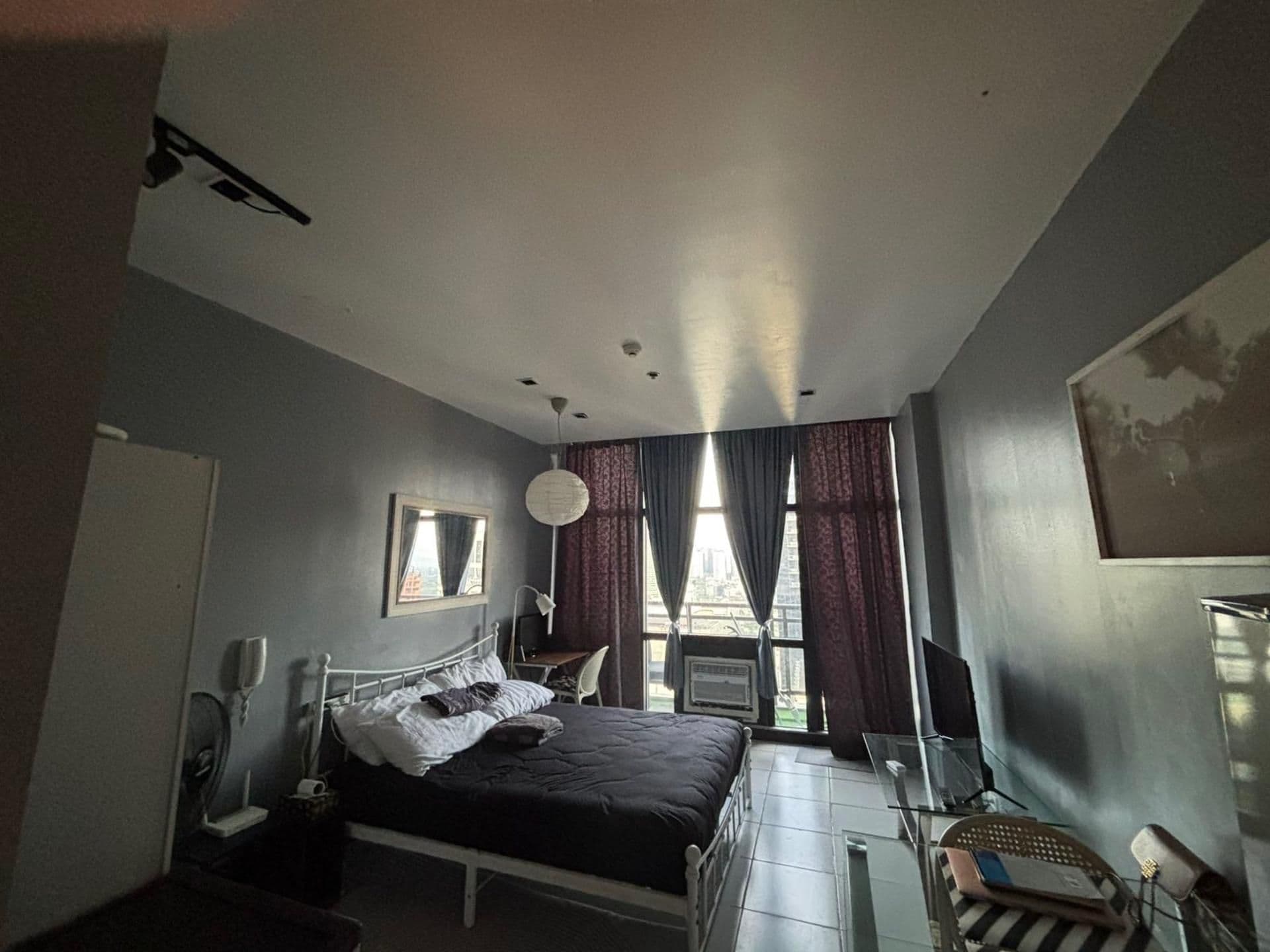 Un dormitorio con cama, escritorio, silla, ventana con cortinas y televisor montado en la pared.