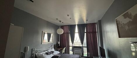 Un dormitorio con cama, escritorio, silla, ventana con cortinas y televisor montado en la pared.