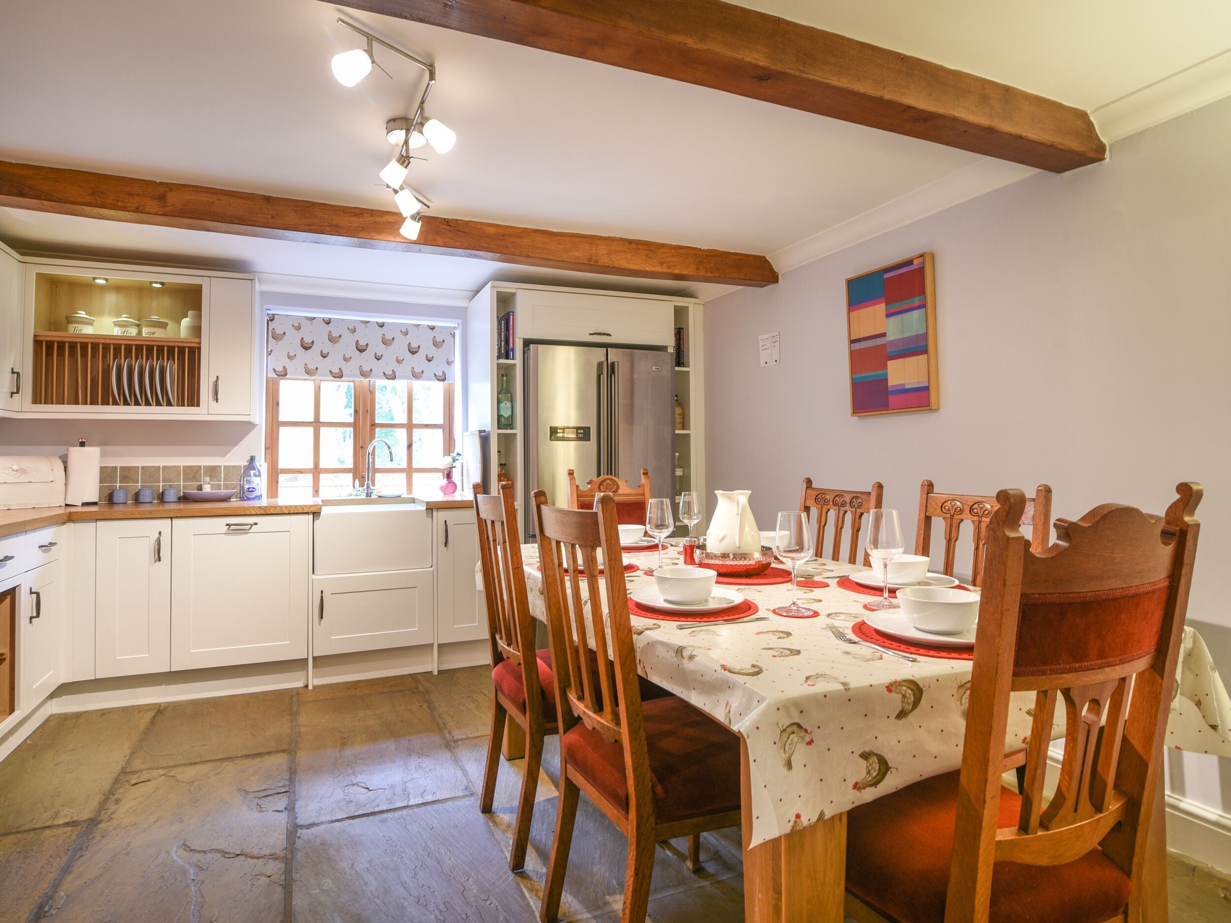 Un comedor con una mesa dispuesta para cuatro personas, rodeada de sillas de madera. Se ve una cocina al fondo con armarios blancos y un ventanal con un motivo de gallinas.
